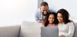 © Prostock-studio - Relaxing Together. Portrait of happy black parents and their cute daughter using laptop, spending time together, watching movie or surfing internet, sitting on sofa in living room, copy space, banner