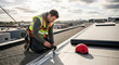 © Владимир Platon - A professional roofer wearing a safety harness and high-visibility vest is on a commercial flat roof, using a tool for repair or installation. The urban skyline is visible in the background