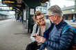 © Marko Geber - Senior couple waiting at train station checking smartphone together