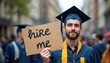 © Vadym - Young graduate holds Hire Me sign in busy street after graduation. Hopeful job seeker faces employment challenges, seeks career opportunity. Displays ambition, determination for pro growth.