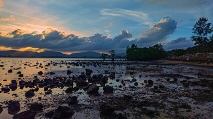 Wall Mural - Time Lapse A dramatic twilight scene of a rocky shoreline during low tide, with silhouetted mountains and trees under a moody, cloud-filled sky. The wet ground reflects the fading light