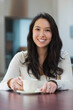 © wavebreak3 - Asian woman smiling while holding white ceramic cup at wooden table in cafe wearing necklace