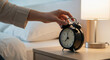 © Vasiliy - Hand reaching for black alarm clock on bedside table in bedroom. Morning routine and time management concept for wake up schedule and daily productivity habits