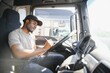 © Serhii - African-American truck driver is sitting behind the wheel holding a clipboard and checking the documents for the cargo