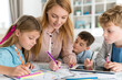© Emvats - Smiling mother helps her children with homework at a table, as the kids draw and write together using colored pencils, with a tablet nearby.