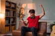 © PaeGAG - Young man raising his fist in the air while holding a smartphone and celebrating a victory in his living room