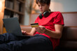 © PaeGAG - Young person completing online transaction, holding credit card while sitting on living room sofa with laptop open