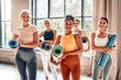 © HBS - A group of diverse, fit women in activewear are holding yoga mats before a workout. They are standing and smiling in a bright, spacious yoga studio.