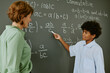 © AnnaStills - Black teenage boy explaining math equation on chalkboard while middle aged Caucasian woman listening attentively in classroom setting, both standing and interacting during lesson
