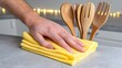 © Finder - Hand Cleaning Surface with Yellow Cloth and Wooden Utensils in Cozy Kitchen Setting with Soft Lighting