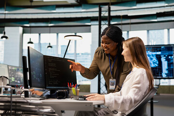 Friendly senior engineer mentoring jolly junior dev at workstation in office, reviewing AI system metrics. Happy african american manager guiding intern through code reviewing using machine learning