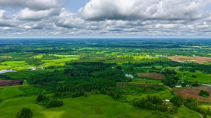 Wall Mural - A beautiful time-lapse captures clouds moving over a scenic Latvian countryside, featuring a winding road, scattered houses, agricultural fields, small lakes, and patches of forest.