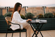 © Darius - Woman works on laptop at outdoor cafe table during sunset on city rooftop