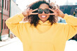 © halayalex - Beautiful woman with afro curls hairstyle.Smiling hipster model in yellow hoodie. Sexy carefree female posing on the street background in sunglasses. Cheerful and happy.Shows peace sign