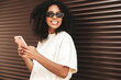 © halayalex - Beautiful woman with afro curls hairstyle.Smiling hipster model in white t-shirt. Sexy carefree female posing in the street near brown wall in sunglasses.Looking at smartphone screen, using apps