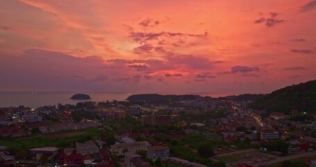 Wall Mural - Aerial view of A breathtaking sunset paints the sky in vivid hues of orange and blue over a coastal town, with lush green hills framing the scene. cloud in dramatic sunset over the ocean