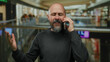 © Krakenimages.com - Man talking on a phone appearing frustrated in an indoor mall with blurred background, beard, bald, and casual attire conveying a sense of urgency and business.