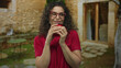 © Krakenimages.com - Woman with curly hair wearing glasses holds red mug at stone building entrance; quiet contentment mindful pause.