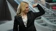 © Krakenimages.com - Young blonde woman in a mall flexing arm power in business attire, showcasing confidence and success against an urban shopping center backdrop with escalators.