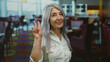 © Krakenimages.com - Senior woman with grey hair making a peace sign in an indoor restaurant setting, exuding positivity and confidence amidst colorful decor and modern interior design.