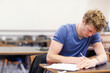 © wavebreak3 - Male student leaning over wooden desk writing on paper in school classroom with pen, copy space