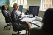 © DragonImages - Caucasian middle aged woman wearing headset sitting at desk in modern office, turning to colleague, while Black young adult woman working at computer in background