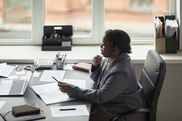 Wall Mural - Black woman sitting at desk wearing headset working on laptop holding pen appearing focused during business call in modern office environment with documents scattered