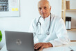 © Andrii Lysenko - old doctor physician using laptop in hospital office. Senior male gp elearning working at computer sitting at desk.