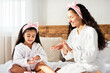 © Prostock-studio - Beautiful young mother and adorable little daughter applying cream on hands, wearing bathrobes and hair bands, sitting on bed at home, copy space. Mom teaching child body care