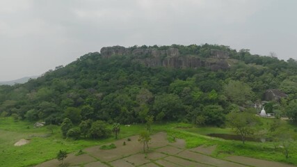 Wall Mural - Drone footage showcasing a green forest hill with rocky cliffs and farmland below under cloudy skies.