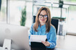 © deagreez - Professional businesswoman in office workspace reviewing documents and smiling while working behind a computer monitor