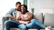 © Prostock-studio - Joyful african american young couple in casual sitting on couch, hugging and laughing, living room interior, copy space. Happy black family spending cold winter day together at home, cuddling