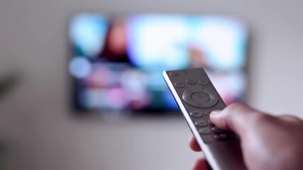 A hand holds a TV remote control, in front of a blurred TV screen showing colorful content