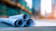 © Curioso.Photography - Rolled architectural blueprints resting on a construction site surface, with scaffolding and buildings visible in the background.