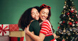 © peopleimages.com - Women, hug and smile in Christmas studio with friends, holiday and festive cheer for bonding. Excited, love and embrace by tree for xmas celebration, presents or decoration on green background