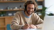 © Sawitree88 - Young, happiness smiling adult student using headphones while taking notes on a laptop at home and participating in an online virtual education class meeting and e-learning webinar.