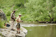© pressmaster - Middle aged man and teenage boy standing on rocky riverbank fishing together with rods, both focused on water, surrounded by green trees and flowing river