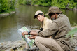 © pressmaster - Middle aged man and teenage boy sitting on riverbank studying map together, father teaching son navigation skills during outdoor fishing trip, lush green trees in background