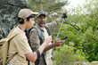 © pressmaster - Middle aged South Asian man showing smartphone to teenage South Asian boy while standing outdoors with backpacks, both holding fishing rods, surrounded by green forest and rocky terrain