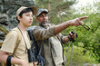 © pressmaster - Middle aged man and teenage boy standing outdoors in forest, man pointing into distance while boy watching attentively, both carrying fishing and hiking gear