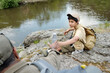 © pressmaster - Middle aged man handing water bottle to boy sitting on rocky riverbank, both wearing outdoor gear, father and son taking break during fishing trip near water