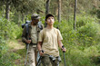 © pressmaster - Middle aged man and teenage boy hiking together in forest, both carrying backpacks and trekking poles, father following son along wooded trail surrounded by green trees