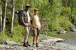 © pressmaster - Middle aged Black man and teenage boy standing together on rocky riverbank holding fishing rods, wearing outdoor gear, surrounded by green trees and flowing water