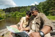 © pressmaster - Middle aged man and teenage boy sitting together outdoors near river studying map, father teaching son fishing techniques in nature, both focused on navigation and planning activity