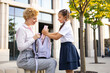 © sofiko14 - A mother helps her daughter pack her backpack in a schoolyard setting.