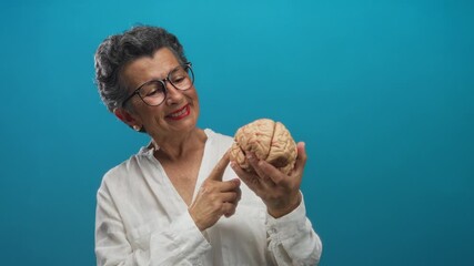 Senior woman with glasses smiling and pointing at a brain model against a vibrant blue background, conveying themes of education and wisdom.