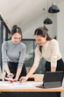 © kenchiro168 - Two Asian women, smiling and collaborating in modern office, review documents and charts on desk with laptop, expressing teamwork and productivity in bright workspace