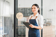 © kenchiro168 - Young Asian woman barista wearing apron holding digital tablet standing in modern coffee shop with open sign, smiling and ready to serve customers