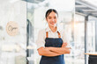 © kenchiro168 - Young Asian woman barista in white shirt and denim apron standing with arms crossed, smiling confidently at modern coffee shop entrance with open sign, welcoming atmosphere