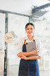 © kenchiro168 - Young Asian woman wearing apron stands in front of glass door with open sign, holding digital tablet and stylus, smiling confidently in modern cafe setting